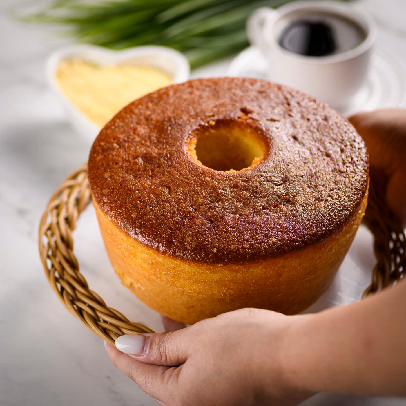 Bolo de fubá inteiro sobre cesta de palha, ao lado de uma xícara de café